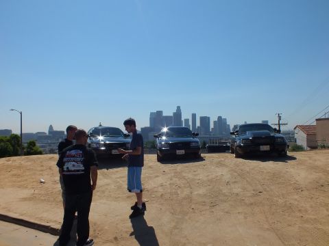2003 Ford Crown Victoria - Black Crown Victorias lined up on a hilltop dirt lot, LA high-rises hazy behind them; two men stand at left.