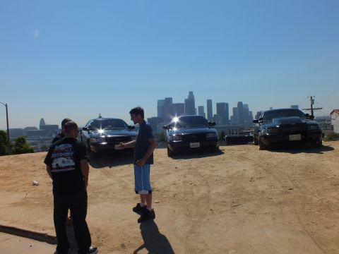 2003 Ford Crown Victoria - man holding keys talks with another in a graphic tee; three black sedans face the LA skyline behind them.