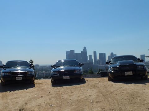 2003 Ford Crown Victoria - Trio of black sedans front-on at a dusty overlook; the right car wears a chrome push bar, downtown LA beyond.