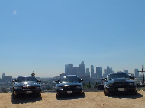 2003 Ford Crown Victoria - Three black sedans spread across a hilltop dirt lot, downtown LA skyline faint in the distance.