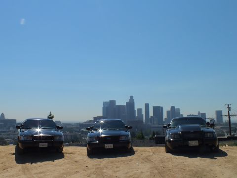 2003 Ford Crown Victoria - Front view of three black sedans on a dusty lot; the right one carries a push bar, LA high-rises behind.