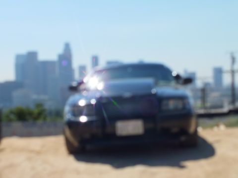 2003 Ford Crown Victoria - Blurred front shot of a black Crown Victoria with push bar; headlights flare against a hazy LA backdrop.