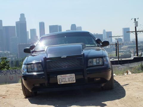 2003 Ford Crown Victoria - Three-quarter front of a black Crown Victoria with push bar and EVIL VIC plate, LA towers behind.