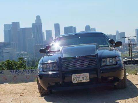 2003 Ford Crown Victoria - Black Crown Victoria front with billet grille and brush guard, plate reads EVTL VTC, LA skyline behind.