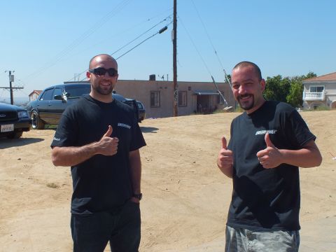 2003 Ford Crown Victoria - Two men in matching black tees give thumbs-up; a black sedan and a stucco house sit behind on the dirt lot.