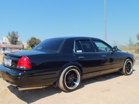 2003 Ford Crown Victoria - Rear three-quarter of a lowered black Crown Victoria on staggered black wheels, parked on a dirt lot.
