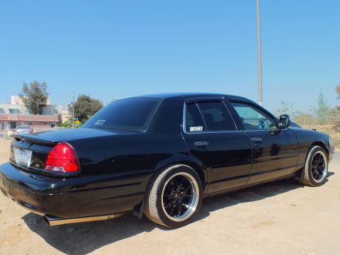 2003 Ford Crown Victoria - Black Crown Victoria from the rear quarter, tinted glass and black multi-spoke wheels, parked on dirt.