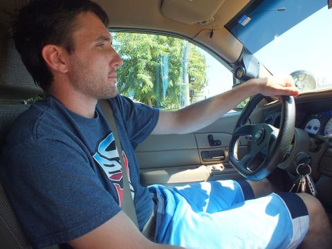 2003 Ford Crown Victoria - man in a graphic tee drives with both hands on the wheel; tan interior and gauge cluster in view.