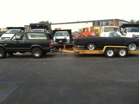 1989 Ford Bronco - A black 1989 Ford Bronco tows a black car on a trailer, with other vehicles and a building in the background.