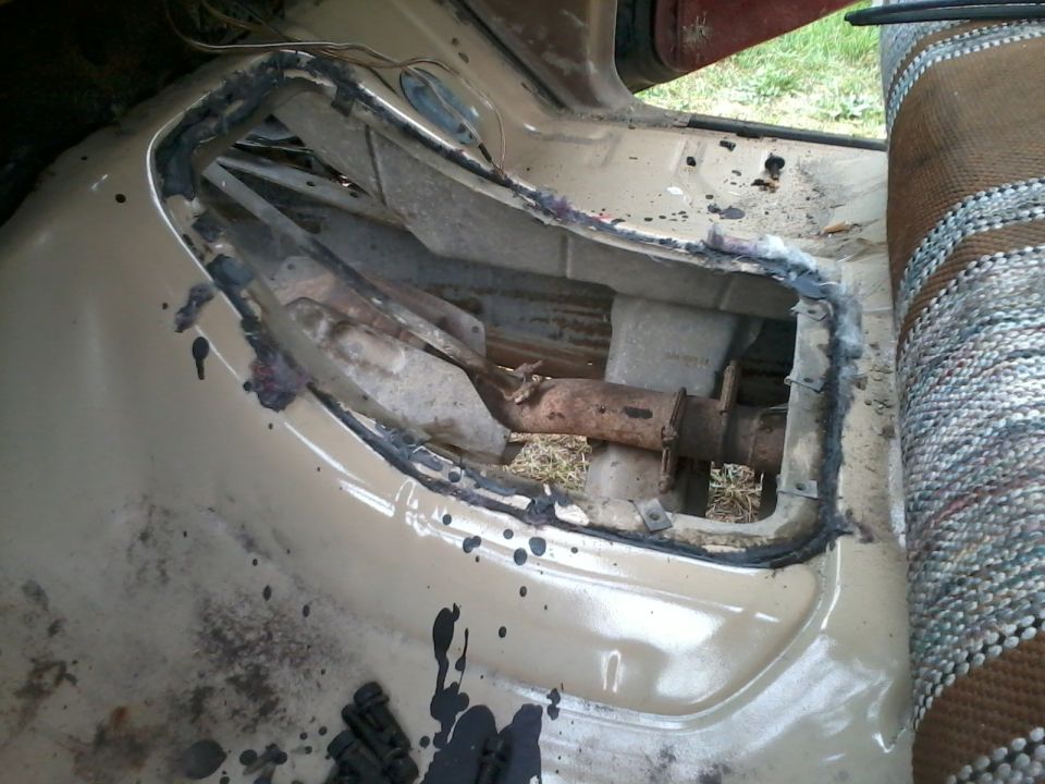 Interior shot of a vehicle's floor pan with a cut-out revealing drivetrain parts underneath. Seat fabric visible on the right