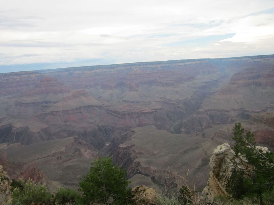 Grand Canyon landscape with rock formations and distant canyon walls under a cloudy sky. Watermark reads SUPERMOTORS