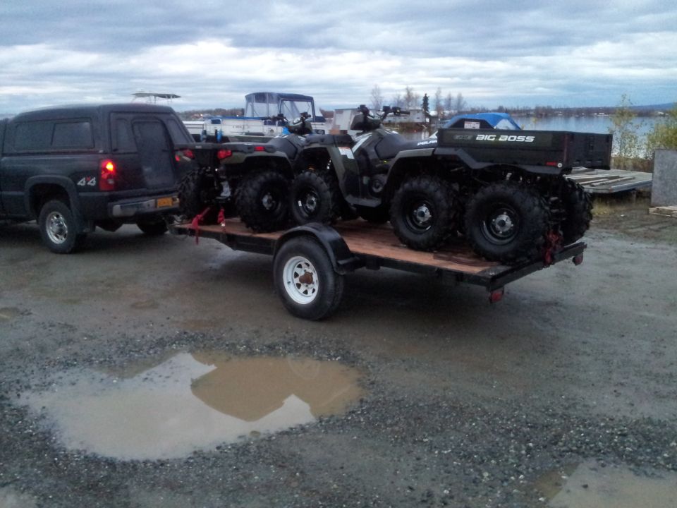 Black pickup truck towing a flatbed trailer with two Polaris ATVs. One ATV has 'BIG BOSS' on the rear cargo box