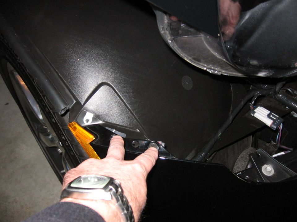 Close-up of a car's black fender and amber turn signal assembly with a hand pointing to a bolt