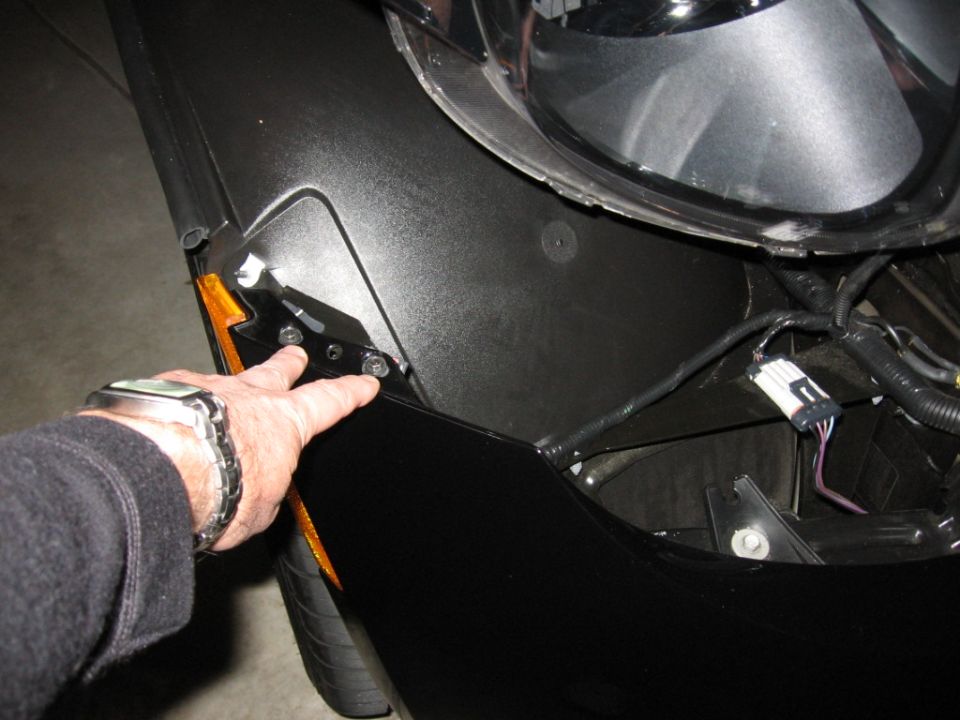 Hand pointing to headlight mounting bolts and wiring harness on a black car