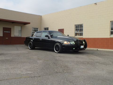 2003 Ford Crown Victoria - Daylight three-quarter shot of the dark Crown Victoria near a CUSTOM MFG CO doorway.