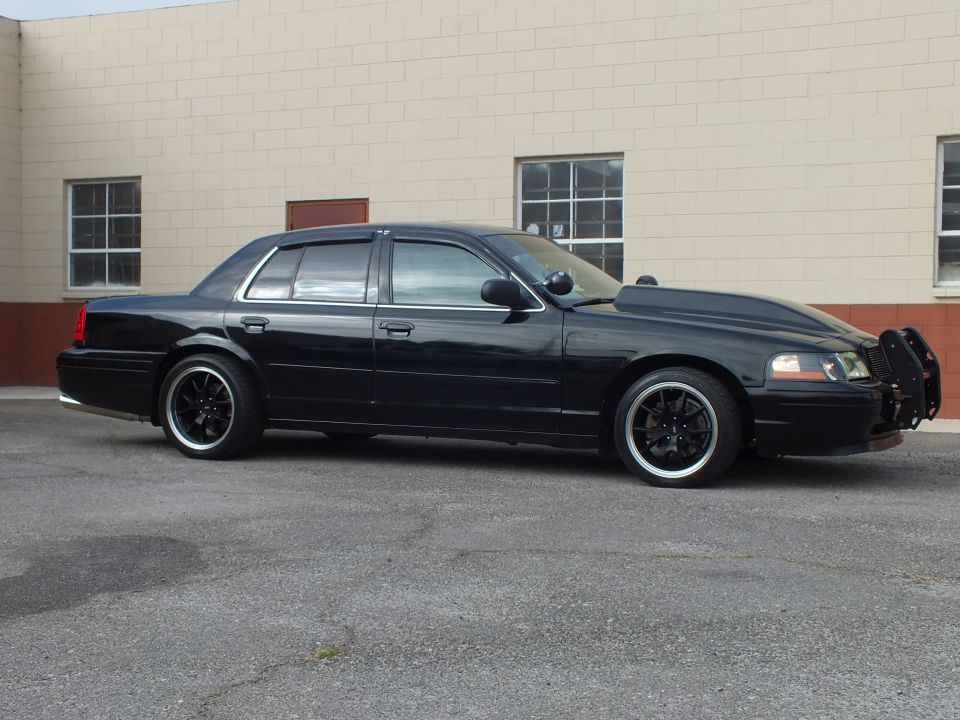 Driver-side profile of black Crown Victoria with grille guard and black wheels in parking lot