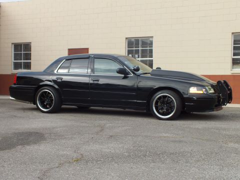 2003 Ford Crown Victoria - Driver-side profile of a blacked-out Crown Victoria parked beside a tan building, black multi-spoke wheels.