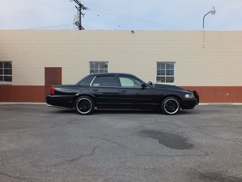 2003 Ford Crown Victoria - Passenger-side profile of a black Crown Victoria beside a brick-and-tan wall, dark tinted windows.