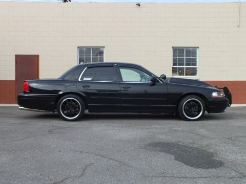 2003 Ford Crown Victoria - Side view of a blacked-out full-size sedan with chrome trim and a front push bar against a brick wall.