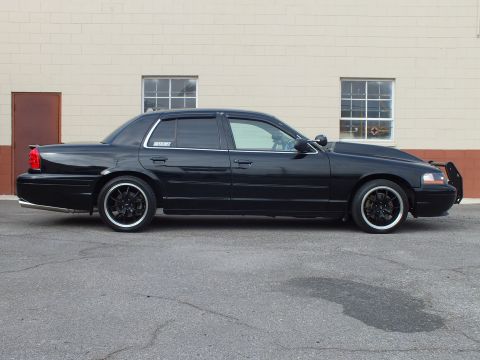 2003 Ford Crown Victoria - Three-quarter passenger profile of a black sedan with bull bar and tinted glass on cracked asphalt.