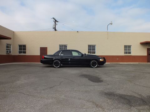 2003 Ford Crown Victoria - Lowered black sedan parked far across a lot in front of a long tan industrial building.