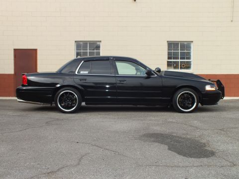 2003 Ford Crown Victoria - Clean passenger profile of a black full-size sedan against a white brick building, dark tint.