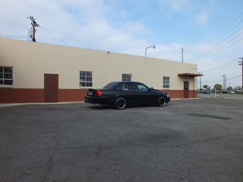 2003 Ford Crown Victoria - Rear driver-side angle of a lowered black sedan near a tan building, with a road and trees behind.