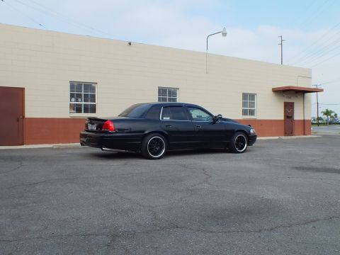2003 Ford Crown Victoria - Rear three-quarter of a black sedan with lip spoiler and tinted windows beside a tan wall.