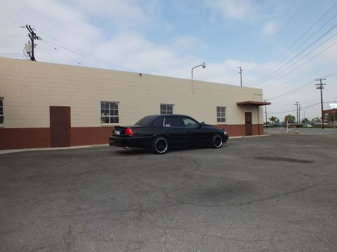 2003 Ford Crown Victoria - Lowered black sedan parked across a wide lot near a tan building and a stop sign.