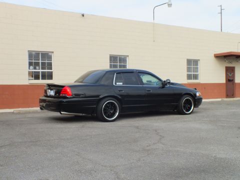 2003 Ford Crown Victoria - Rear corner view of a black sedan with trunk spoiler and tinted glass beside a tan building.