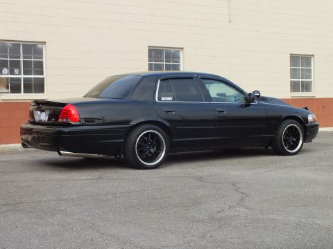 2003 Ford Crown Victoria - Rear three-quarter of a blacked-out full-size sedan with lip spoiler against a brick wall.