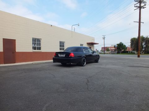 2003 Ford Crown Victoria - Lowered black sedan parked at the corner of a tan building with a street and trees behind.