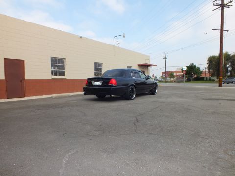 2003 Ford Crown Victoria - Lowered black sedan parked across a wide lot beside a tan building with traffic in the distance.