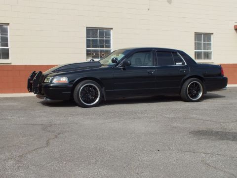2003 Ford Crown Victoria - Murdered-out Crown Victoria parked by a tan building, front push bar and blacked-out trim visible.