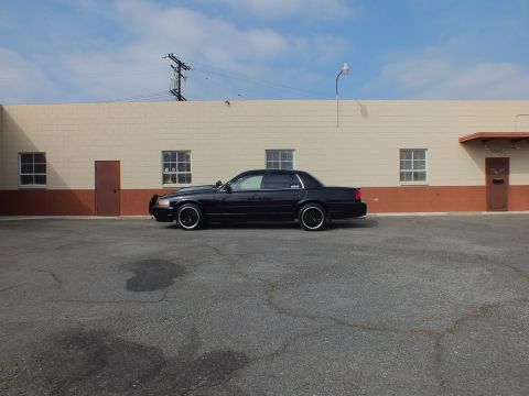 2003 Ford Crown Victoria - Wide shot of a black Crown Victoria sedan in an empty lot beside a warehouse building.