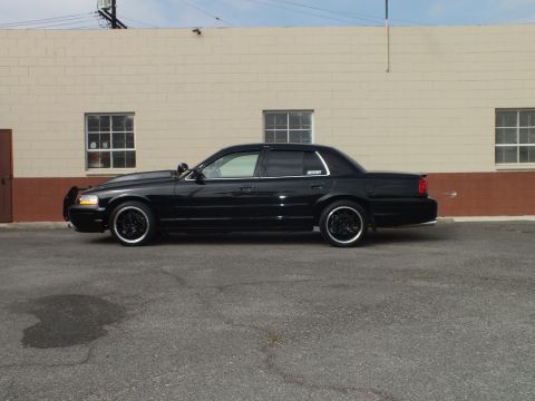 2003 Ford Crown Victoria - Side profile of a murdered-out Crown Victoria parked on asphalt near a tan masonry wall.