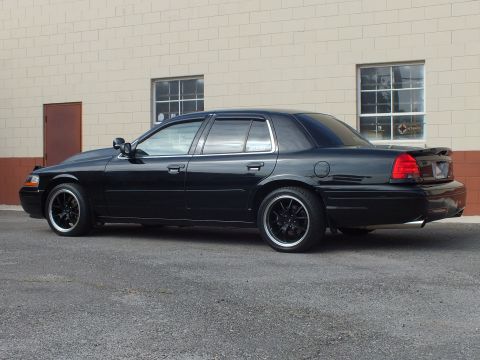 2003 Ford Crown Victoria - Glossy black Crown Victoria rear quarter with window visors and lip spoiler by brick wall.