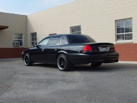 2003 Ford Crown Victoria - Rear quarter of a glossy black Crown Victoria with window visors near a masonry wall.