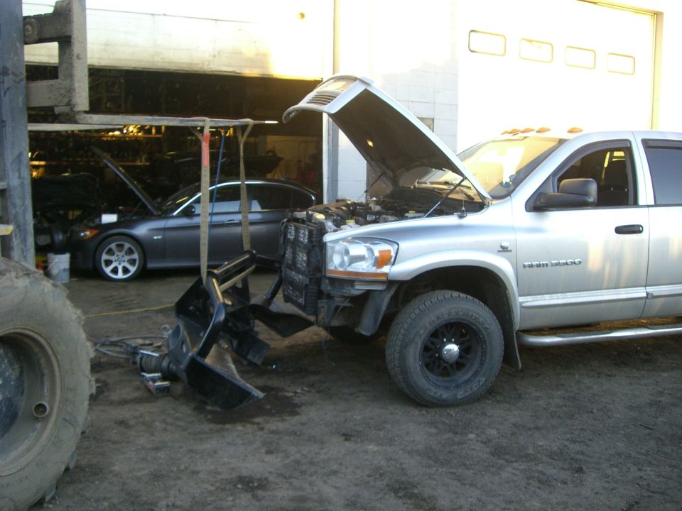 Silver Dodge pickup truck, front bumper removed, hood open, parked in garage. Another car is visible in the background