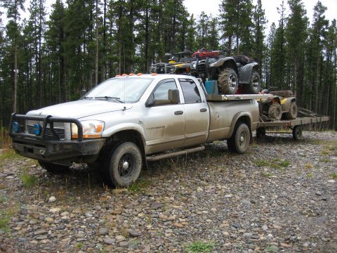 2005 Dodge 3500 - A silver pickup truck carries ATVs on its flatbed and tows a trailer with an additional ATV, parked on a gravelly surface near a forest.