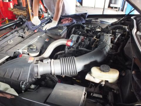 2003 Ford Crown Victoria - Open engine bay showing a wide intake tube and reservoir while a gloved worker reaches inside.