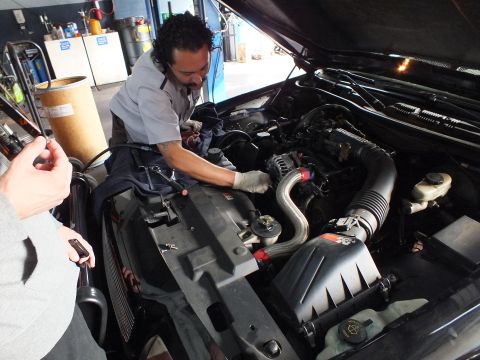 2003 Ford Crown Victoria - Technician reaches into an open engine bay, hands near the intake tube and alternator, shop bays visible behind.