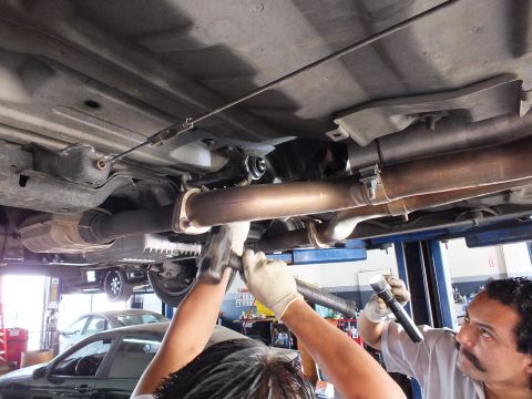 2003 Ford Crown Victoria - Two workers in gloves handle a section of exhaust pipe beneath a raised vehicle; shop floor and cars in background.
