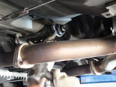 2003 Ford Crown Victoria - Gloved hand steadies a polished driveshaft tube running alongside exhaust components under a lifted car.