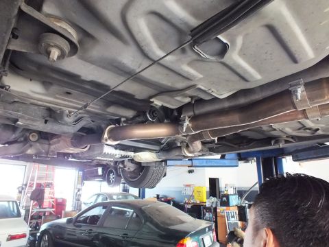 2003 Ford Crown Victoria - Underbody shot of exhaust routing and crossmember; a worker and a dark green sedan appear in the shop behind.