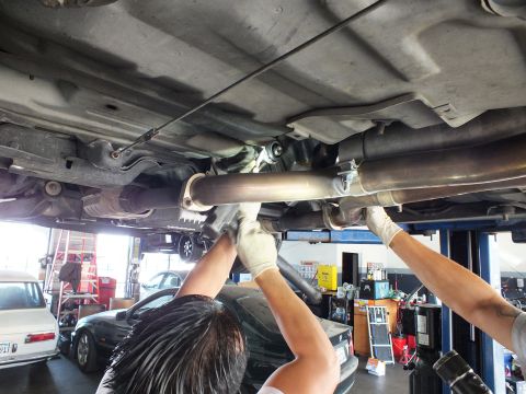2003 Ford Crown Victoria - Workers in gloves push a driveshaft and exhaust section up against the chassis of a lifted car; bays behind.