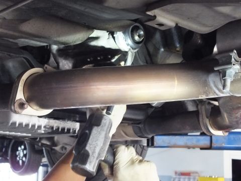2003 Ford Crown Victoria - Gloved hand grips a hammer below a driveshaft tube near the transmission output seal and exhaust components.