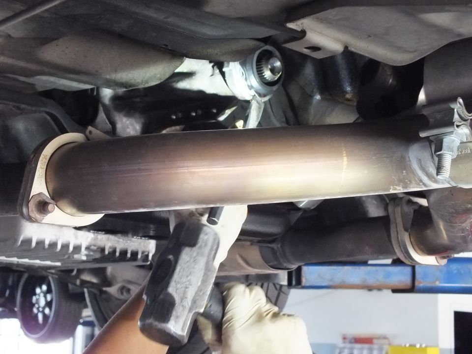 Close-up of driveshaft tube and output shaft seal under vehicle with gloved hand holding hammer