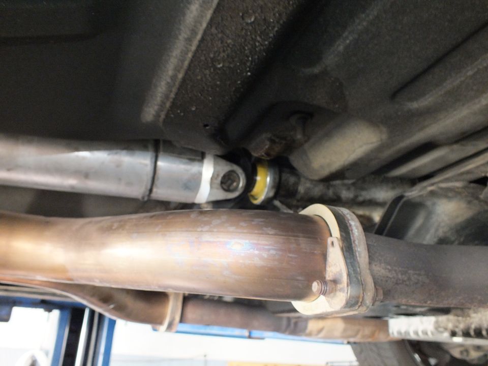 Driveshaft U-joint and yellow carrier bearing alongside polished exhaust pipe underneath a vehicle