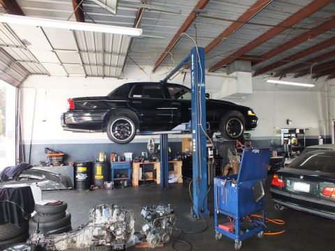 2003 Ford Crown Victoria - Black sedan on a blue two-post lift; a spare engine and parts sit on the shop floor below.